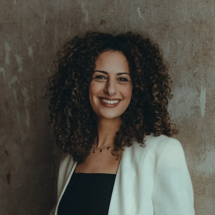 Jasmin lächelt in die Kamera. Sie hat lange braune Locken und trägt ein schwarzes Shirt und einen weißen Blazer.