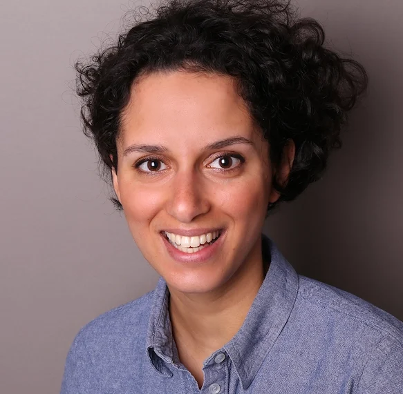 Esther lächelt in die Kamera. Sie hat lange dunkle Locken und trägt ein hellblaues Hemd.