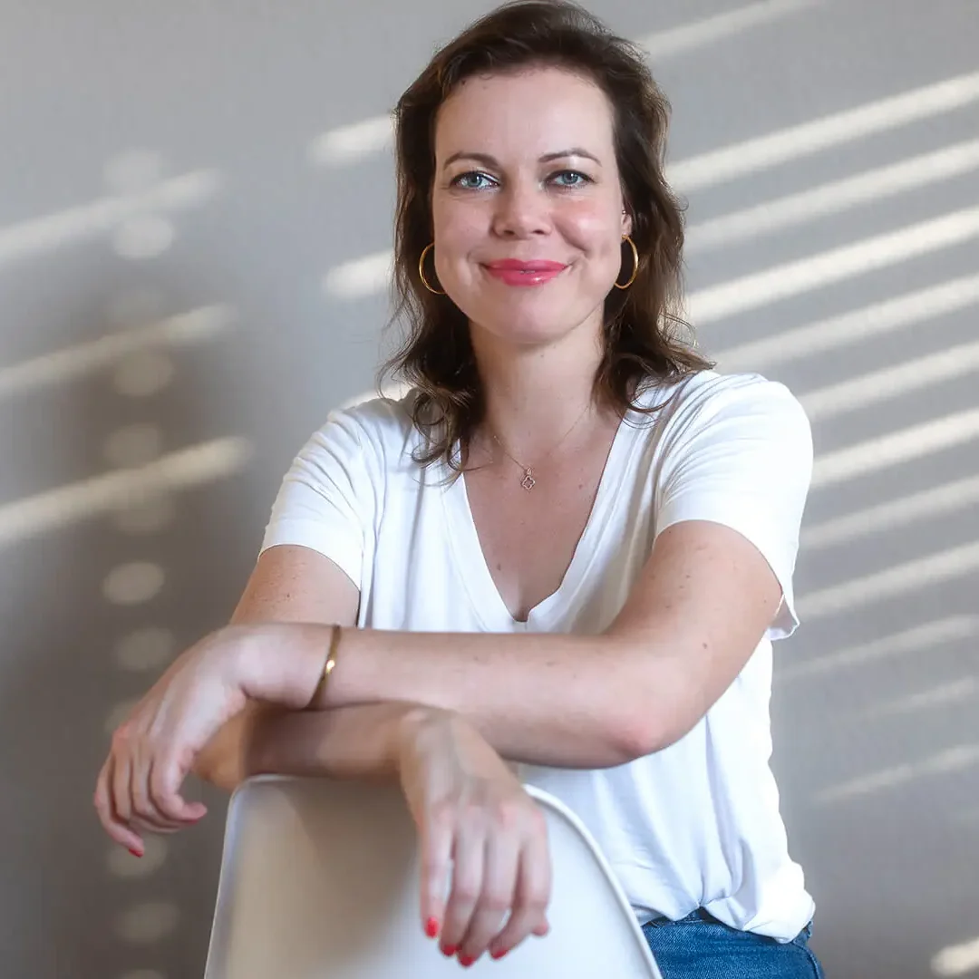 Eva sitzt verkehrt herum auf einem Stuhl, ihre Arme auf die Lehne gestützt und lächelt in die Kamera. Sie hat lange, braune Haare und trägt ein weißes T-Shirt.