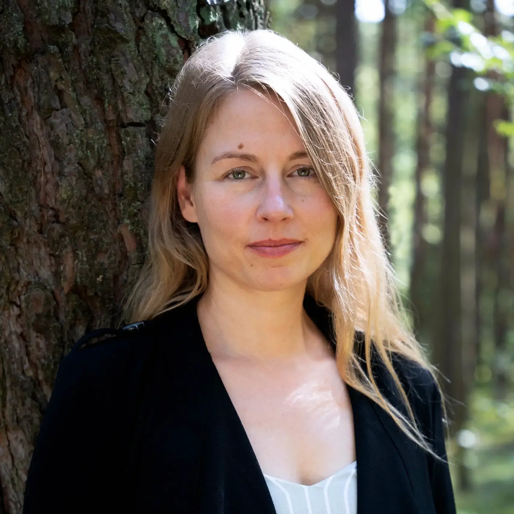 Clara lächelt leicht in die Kamera. Sie hat lange blonde Haare, trägt einen schwarzen Blazer und lehnt mit dem Rücken an einem Baum.