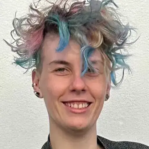 Maike lächelt in die Kamera. Dey hat bunte, verwuschelte Haare und trägt mehrere Ohrringe.