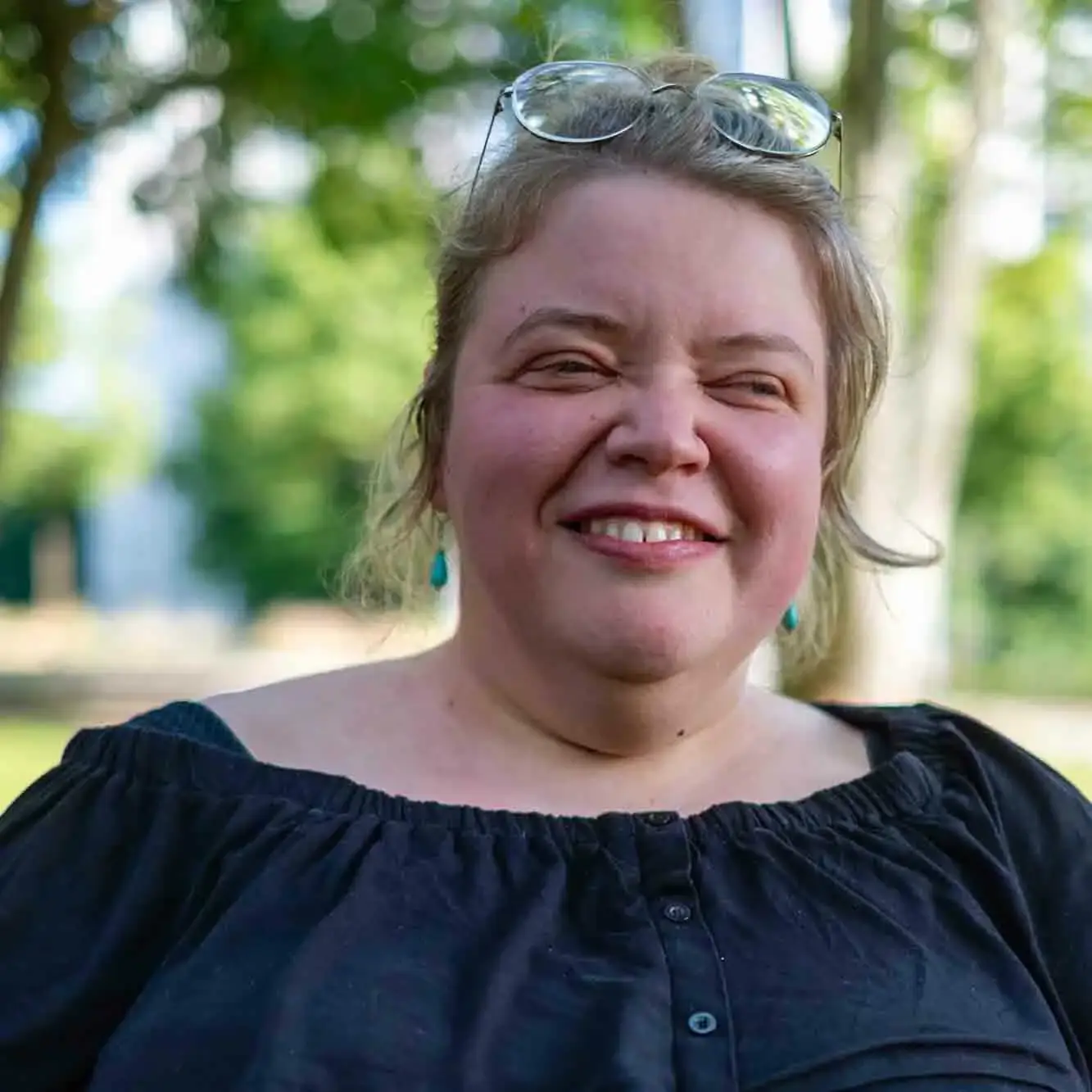 Saskia lächelt in die Kamera. Ihre blonden Haare sind locker zurückgebunden und ihre runde Brille hat sie sich hoch in die Haare geschoben.