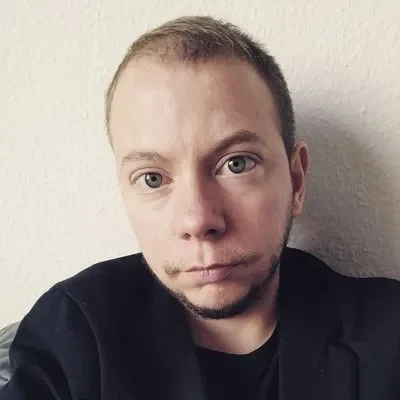 Julien schaut in die Kamera. Er trägt einen schwarzen Pullover und hat kurze dunkelblonde Haare und Bart