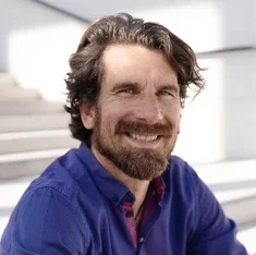 Oliver schaut lächelnd in die Kamera. Er trägt ein violettes Hemd und hat mittellange braune Haare und Bart.