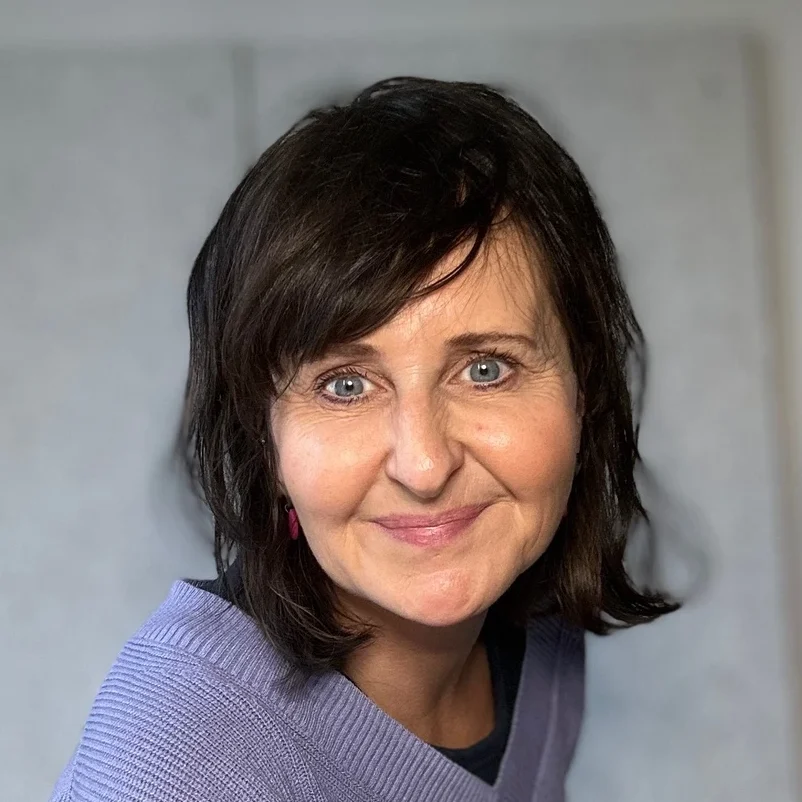 Sarah schaut lächelnd in die Kamera. Sie hat kinnlange schwarze Haare und trägt einen lavendelfarbenen Pullover.