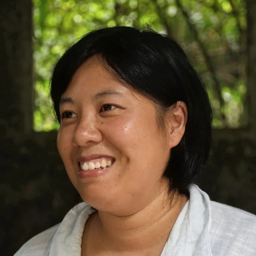 Trang schaut lächelnd an der Kamera vorbei. Sie hat kinnlange schwarze Haare und trägt ein weißes hemd aus Leinen.
