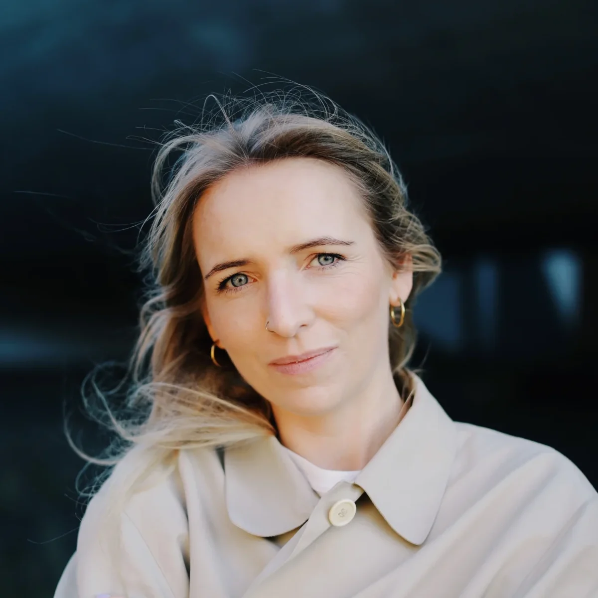 Etienette schaut freundlich in die Kamera. Sie hat lange blode Haare, trägt einen hellen Trenchcoat und hat die Arme verschränkt.